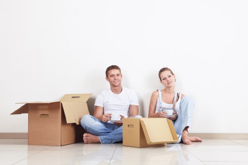 A young couple sitting on a light-colored tiled floor in a residential interior, surrounded by several open and closed cardboard moving boxes of varying sizes, some with flaps open revealing contents, others sealed. The man on the left, dressed in a white t-shirt and blue jeans, is smiling and holding a mobile phone, while the woman on the right, wearing a white top with a light blue cardigan draped over her shoulders, is also smiling and holding a piece of paper. Behind them, more cardboard boxes are stacked against a plain white wall, suggesting an active household move typical of home removals services in Ruislip or nearby areas. The scene is well-lit with natural ambient light, indicating daytime, and the setting appears to be a part of a house or apartment ready for moving or packing, underscoring the context of professional house removals and relocation activities.
