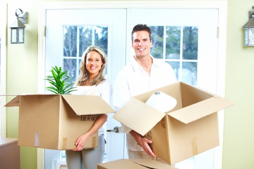 A smiling couple, a woman and a man, are standing in front of a bright, well-lit living room with large glass doors and white frames. They are holding large open cardboard moving boxes, with the woman’s box containing a green houseplant with long, narrow leaves and the man's box partially filled with packing paper and other household items. The scene suggests an ongoing house move, with the individuals prepared to transport their belongings. The room features pale green walls and exterior views of trees through the doors. The presence of the boxes and the cheerful expressions indicate a professional house removals or moving service, consistent with Ruislip Man with Van’s offerings in the local Ruislip area, close to the HA postcode district. The natural lighting from the outdoors highlights the domestic setting, emphasizing an organized and friendly move preparation within a residential property environment.